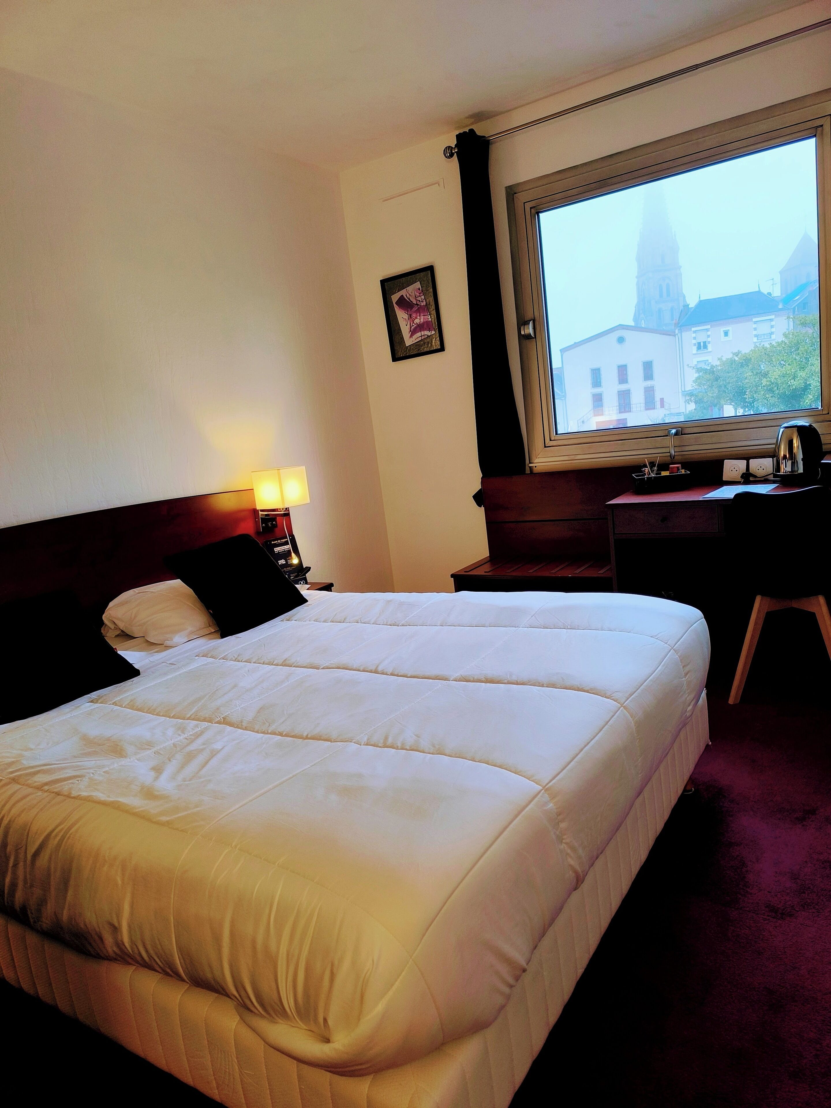 Quarto de hotel com uma cama grande, uma escrivaninha com cadeira, uma janela com vista para um edifício e um quadro pendurado na parede.