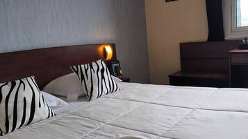 Een net opgemaakt bed met witte beddengoed en kussens met een zebraprint, een nachtkastje met een lamp en een raam met gordijnen.