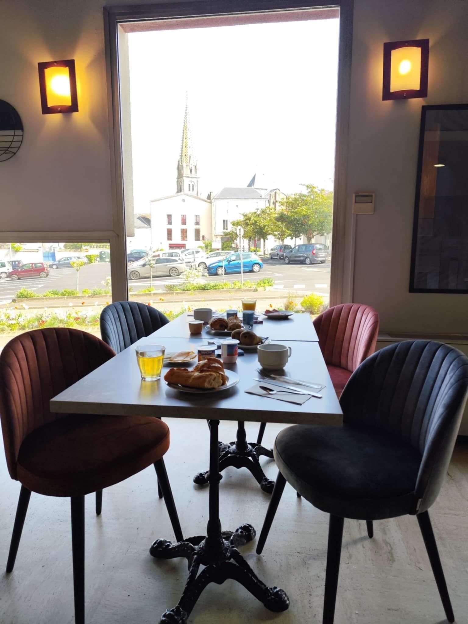 A dining area with a table set for two, chairs, and a view of a street with parked cars and a building with a tower.