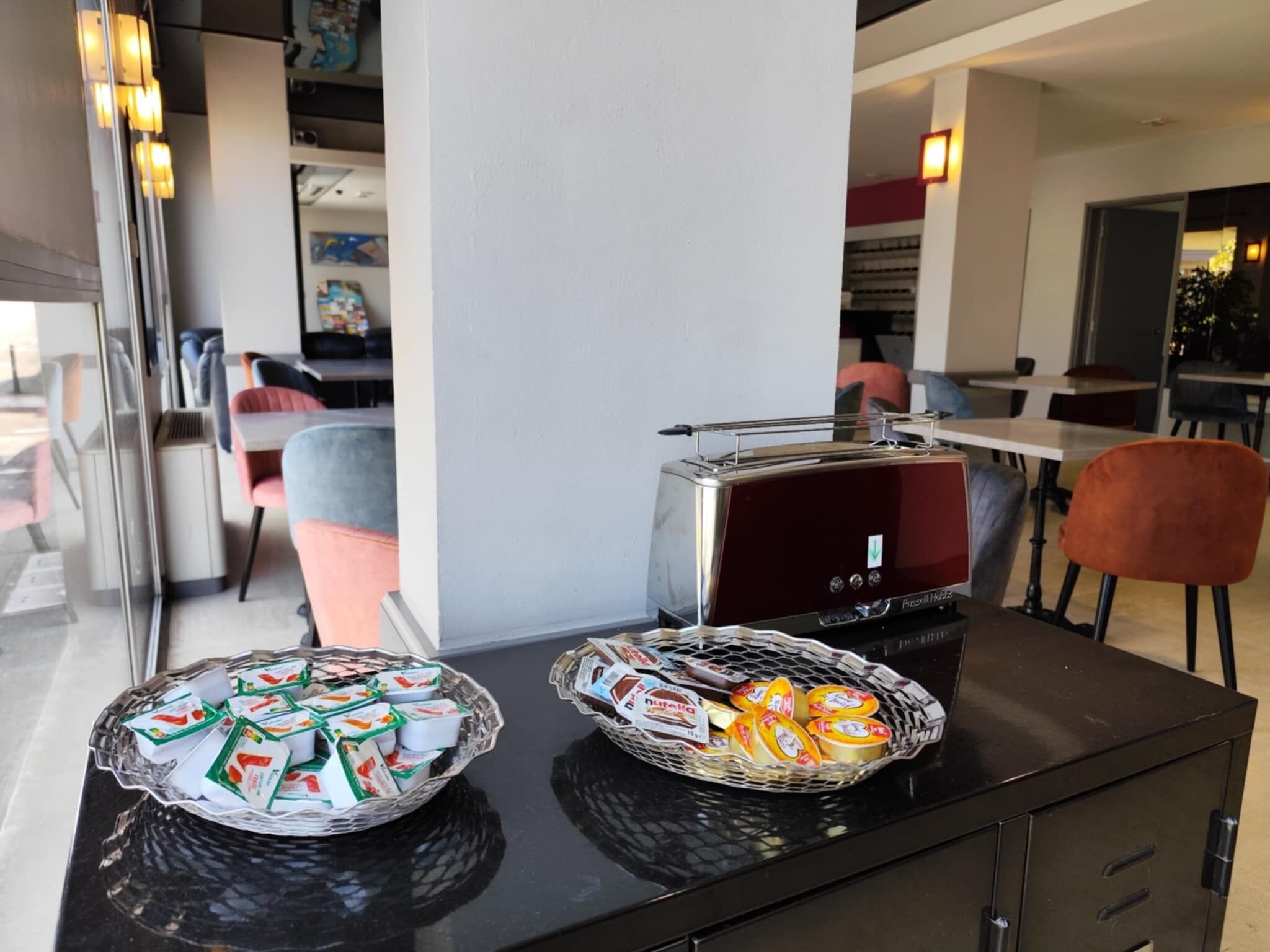 Un buffet de petit-déjeuner avec des yaourts et du Nutella présentés dans des paniers, un grille-pain et un intérieur moderne agrémenté de chaises et de tables.