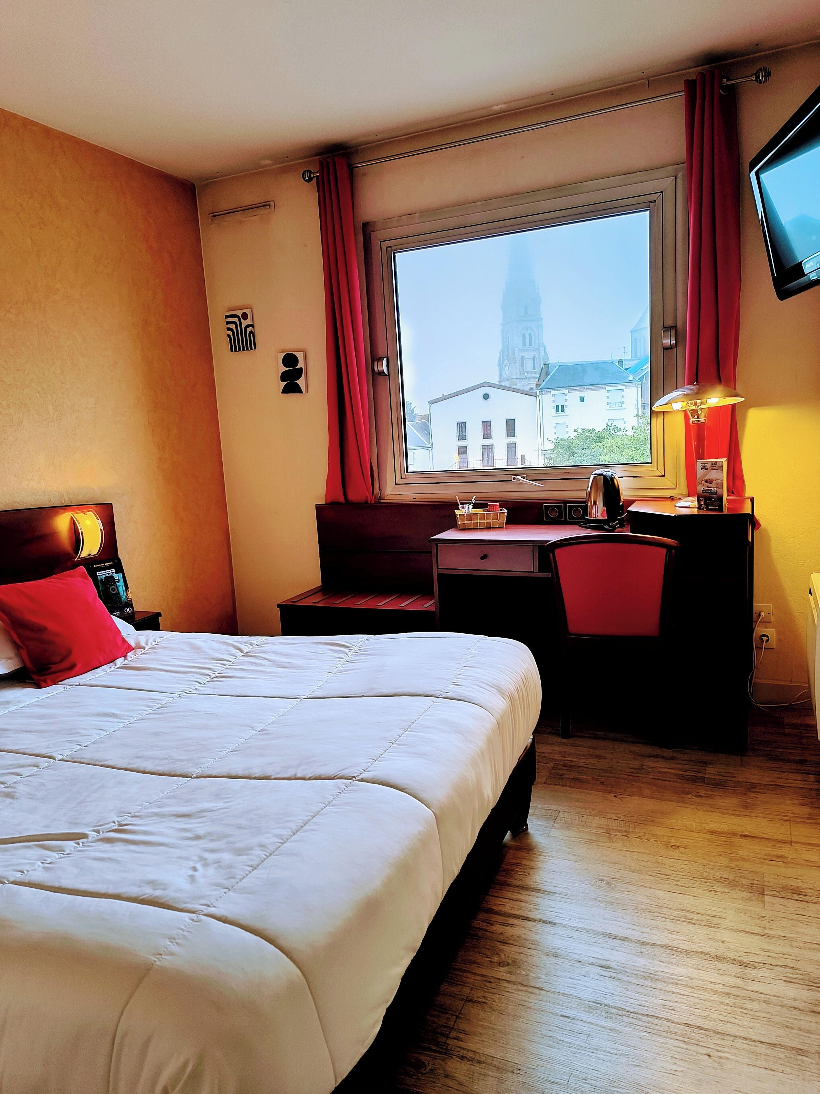 Ein Hotelzimmer mit einem großen Bett, einem Schreibtisch mit Stuhl, einem Fenster mit Blick auf ein Gebäude und einem an der Wand befestigten Fernseher.