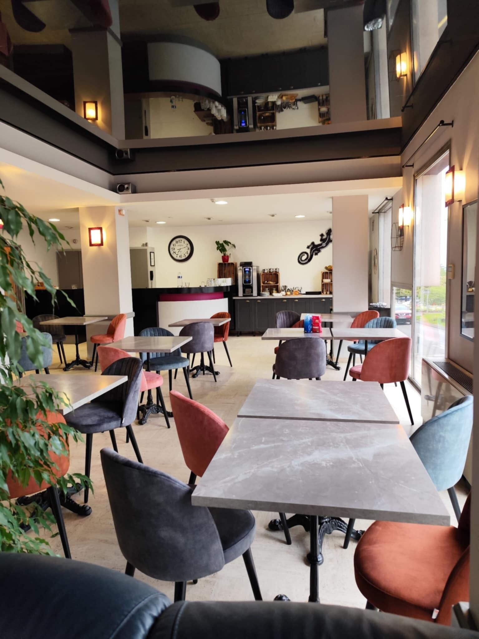 Un comedor interior moderno con varias mesas y sillas, una zona de cocina al fondo y grandes ventanales que aportan luz natural.