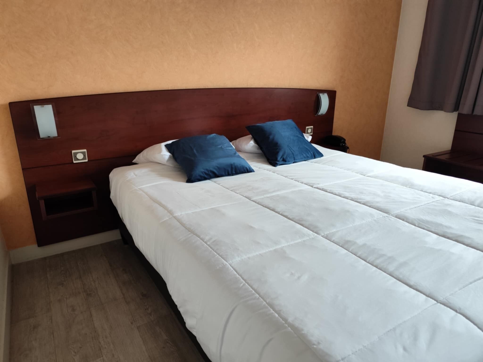 A neatly made bed with white bedding and two blue pillows, a wooden headboard, and a nightstand with a lamp.
