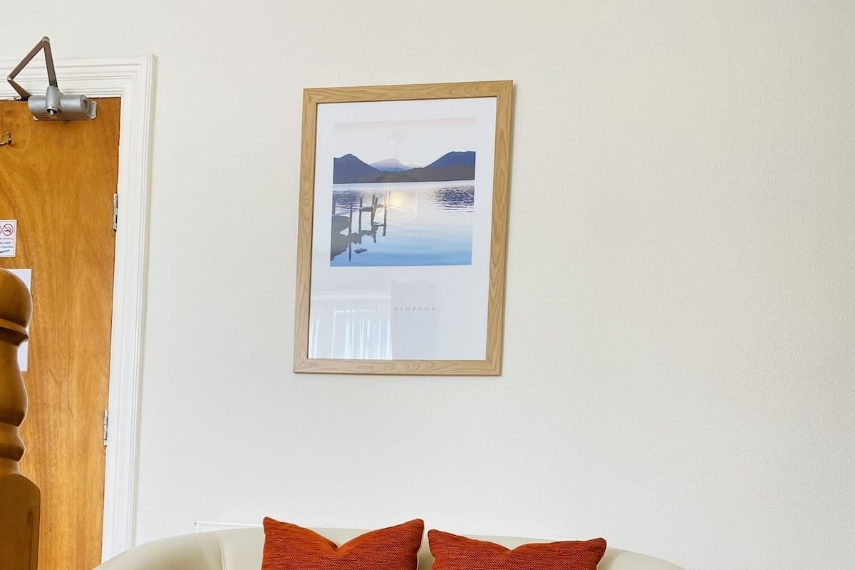 A cream-colored leather sofa with two orange pillows against a white wall, a framed picture above, and a wooden door to the left.