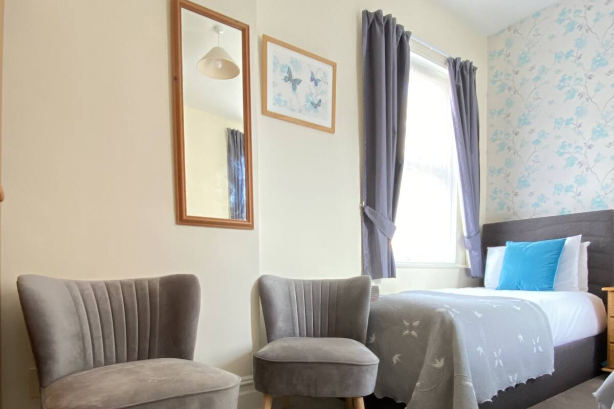 A bedroom with two grey upholstered chairs, a bed with a grey blanket, a nightstand, a mirror, and a window with curtains.