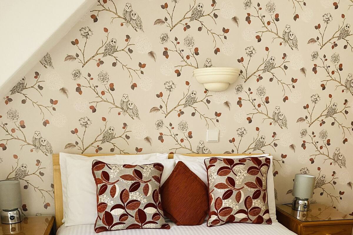 A neatly made bed with patterned pillows, a bedside table with a lamp, and a wall with a floral wallpaper design.