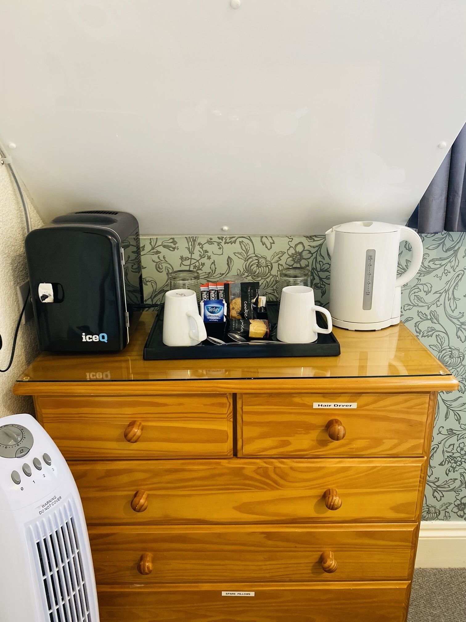 A wooden dresser with a hair dryer, a portable heater, and a tray with coffee mugs, a tea bag, and a box of tea.