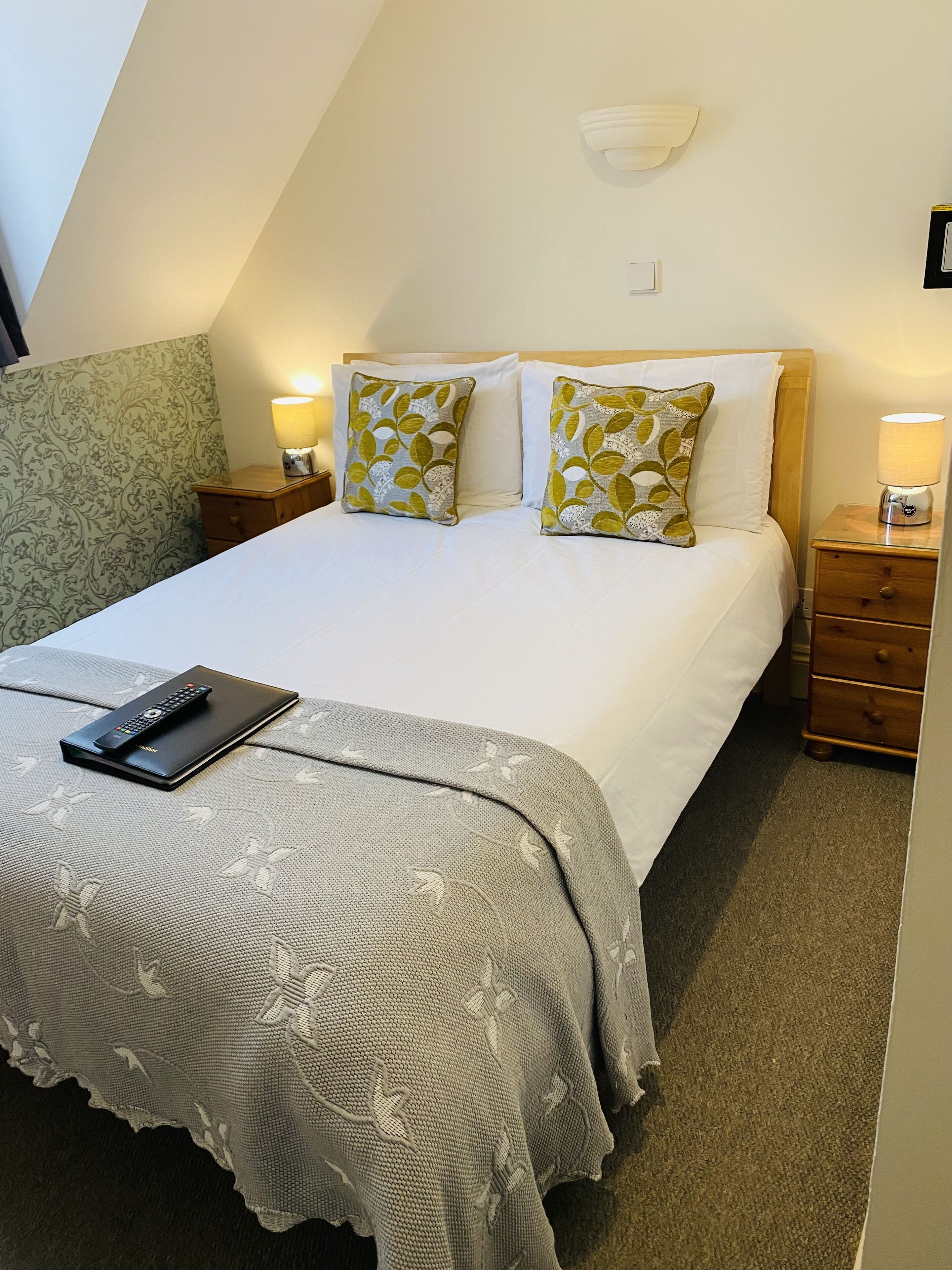 A neatly made bed with pillows, a bedside lamp, a remote control on a blanket, and a patterned wall.