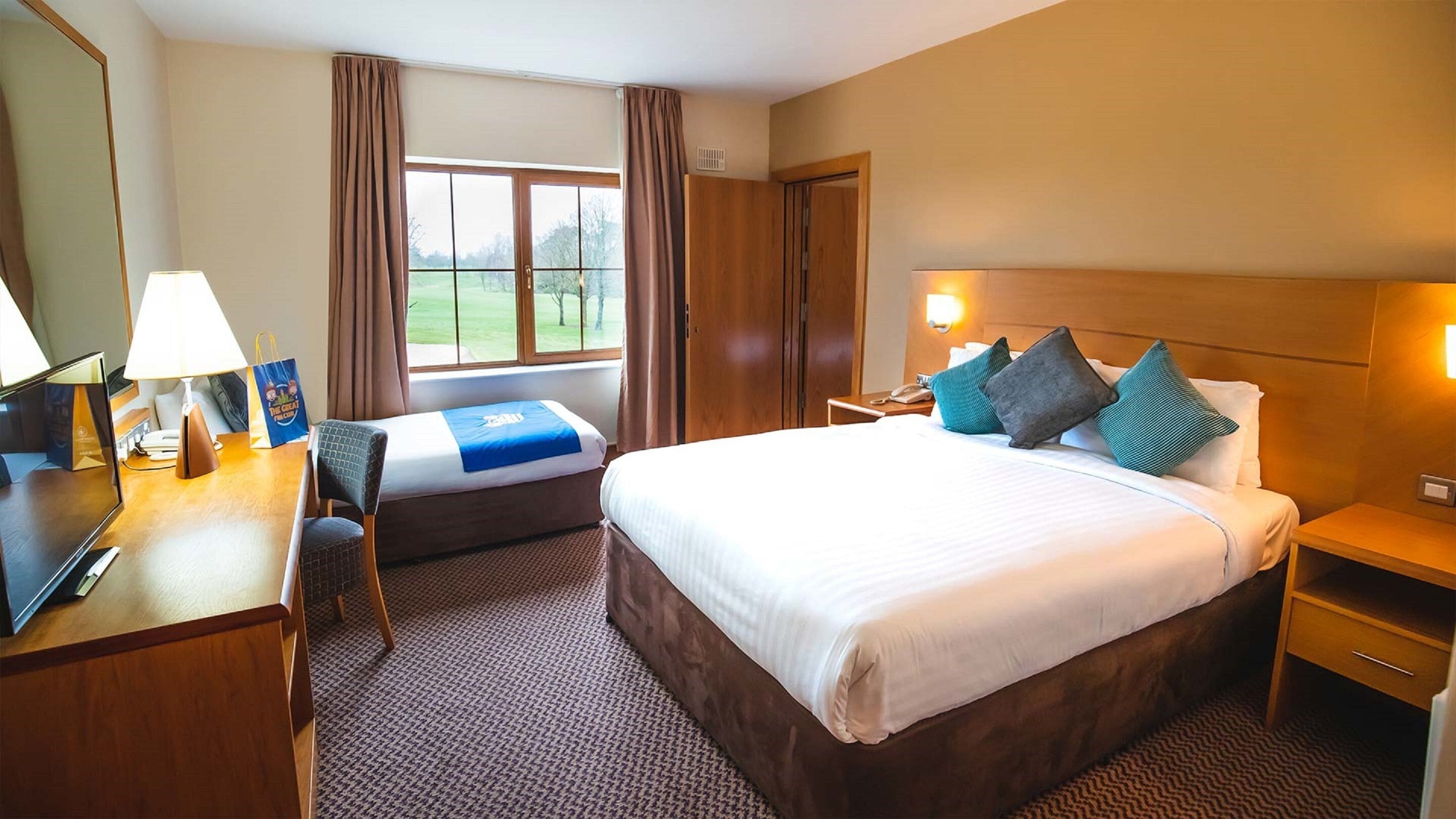 A hotel room with a large bed, a desk with a lamp, a chair, and a view of a green landscape through the window.