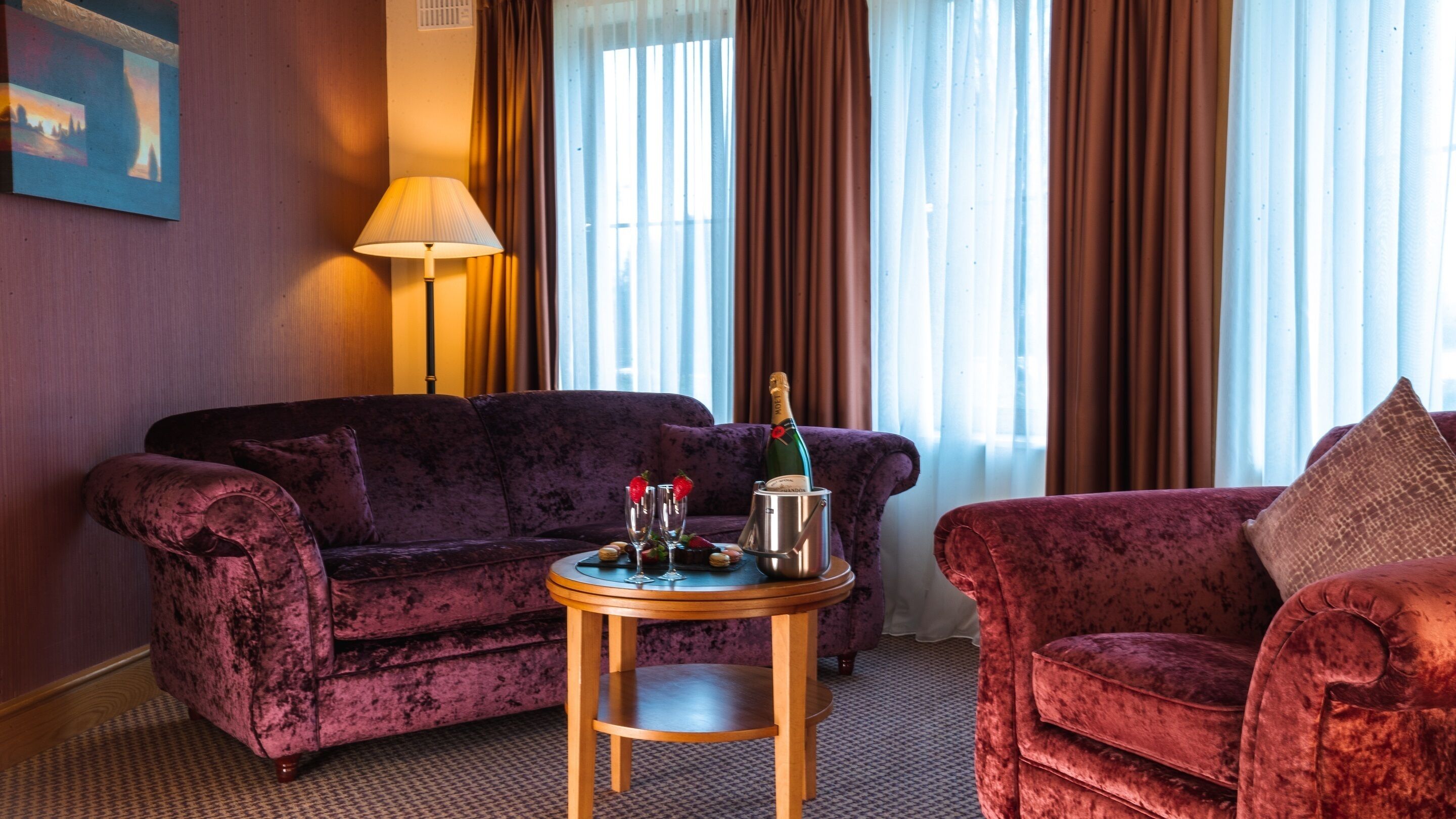 A room with a purple velvet sofa, a wooden side table with a bottle of champagne, glasses, and strawberries, and a red velvet armchair.
