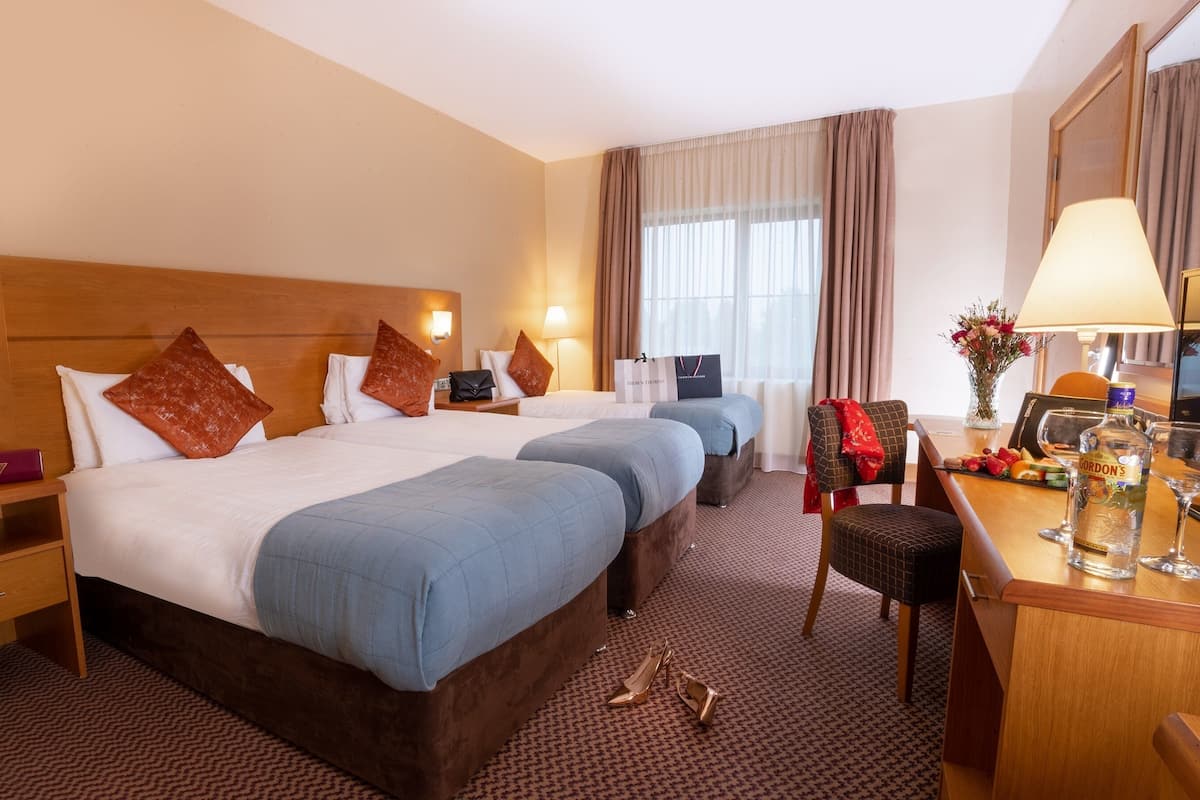 A hotel room with two beds, a desk with a lamp, a chair, a small table with a bottle and glasses, and a window with curtains.