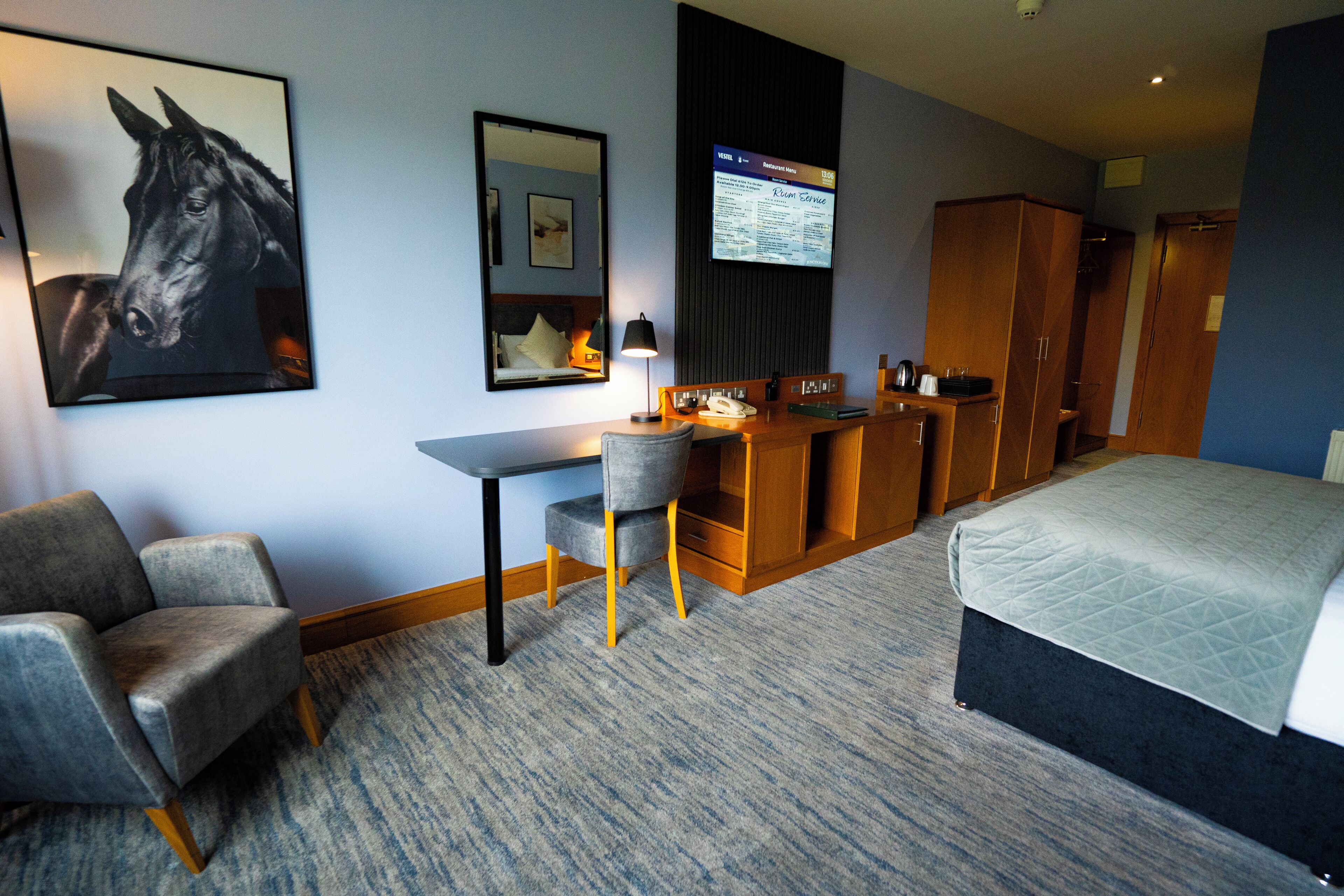 Habitación de hotel con cama, escritorio con silla, armario, televisor montado y retrato enmarcado de un caballo.