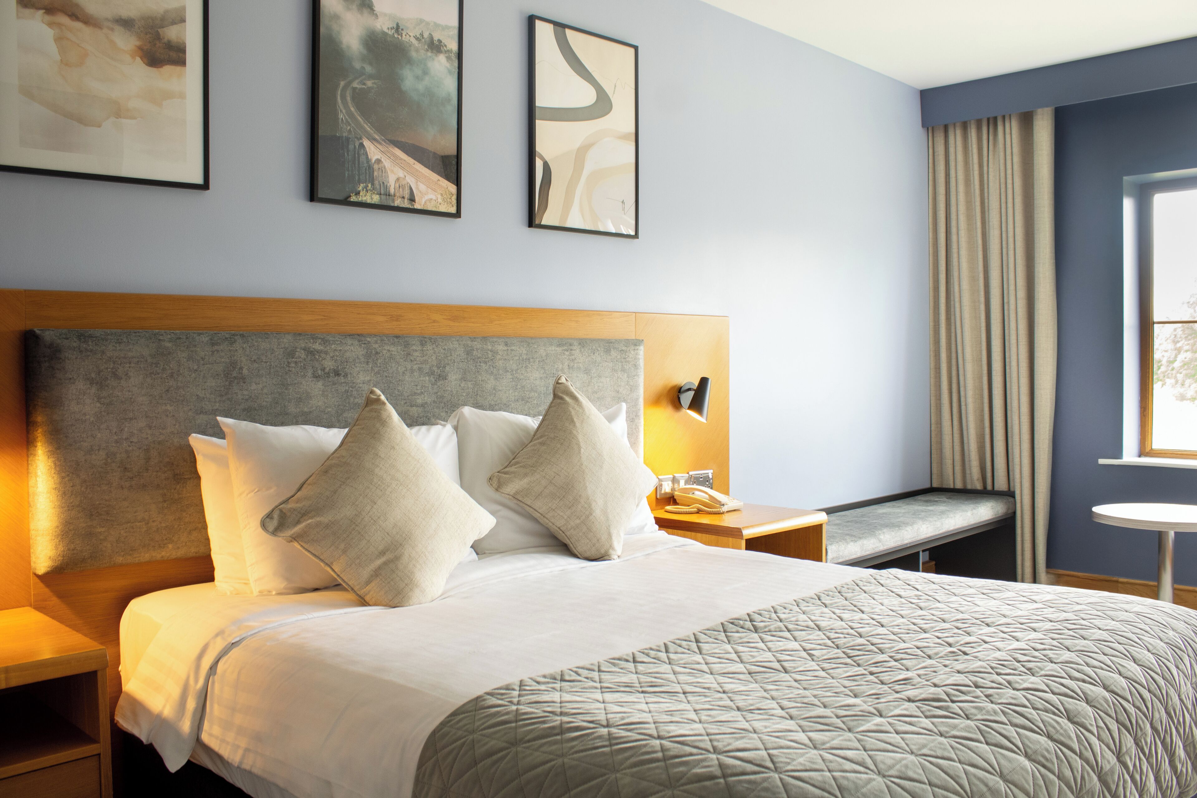 Habitación de hotel con cama, mesita de noche, lámpara, ventana con cortinas y una obra de arte en la pared.