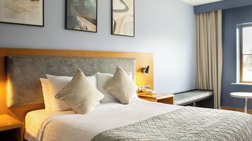 Habitación de hotel con cama, mesita de noche, lámpara, ventana con cortinas y una obra de arte en la pared.