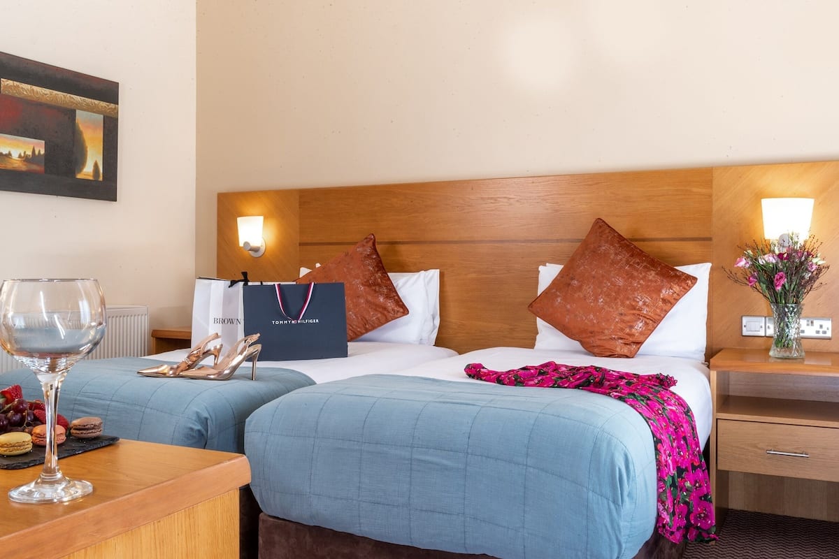 A hotel room with two beds, a wooden headboard, a nightstand with a vase of flowers, and a table with wine glasses and snacks.