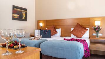 A hotel room with two beds, a wooden headboard, a nightstand with a vase of flowers, and a table with wine glasses and snacks.
