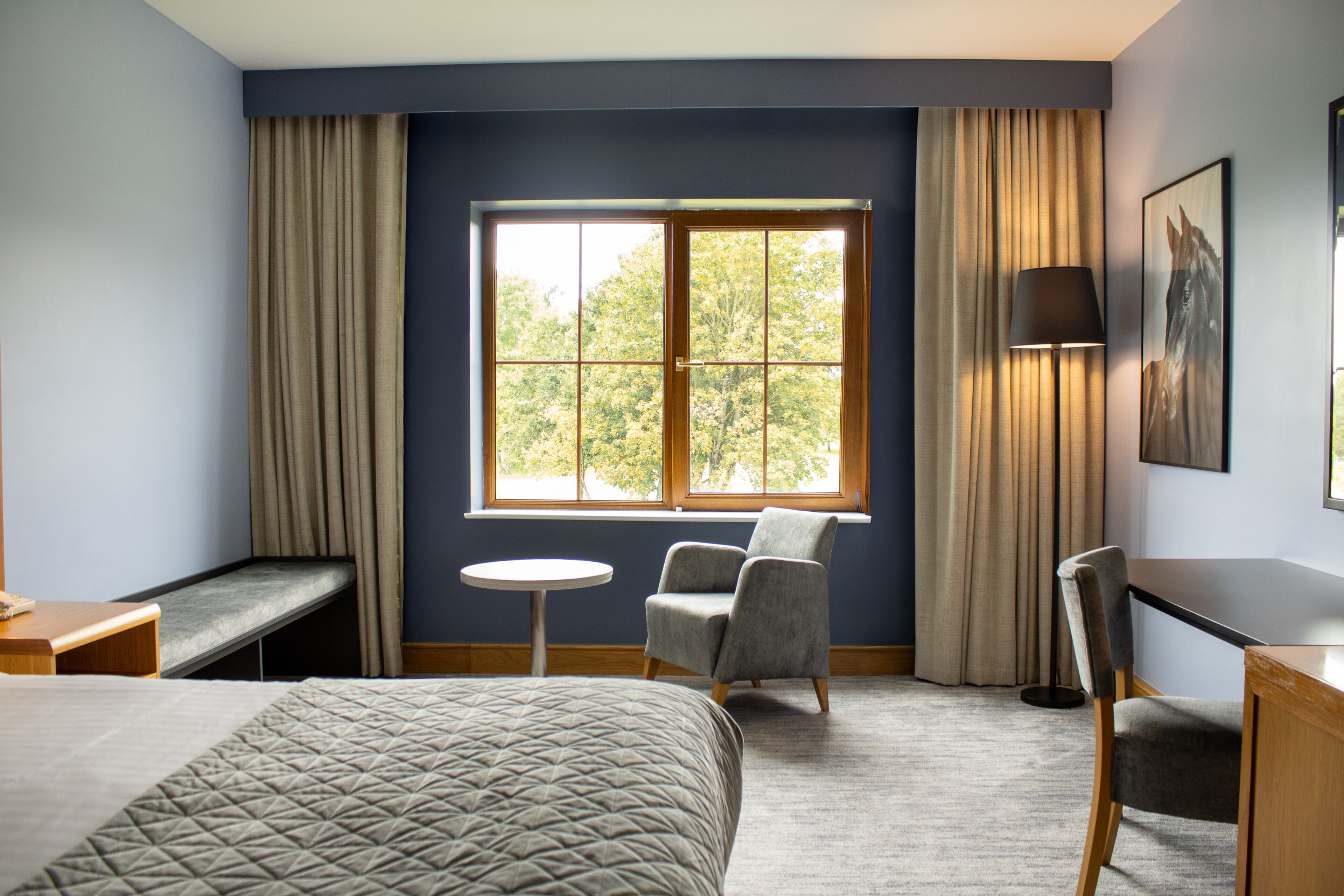 A hotel room with a bed, two chairs, a small table, a lamp, and a window with curtains.