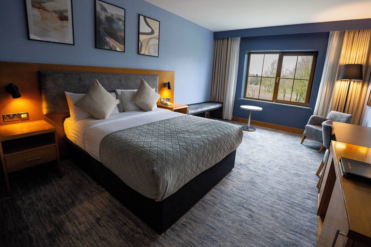 A hotel room with a large bed, a desk, a chair, a small table, and a window with curtains.