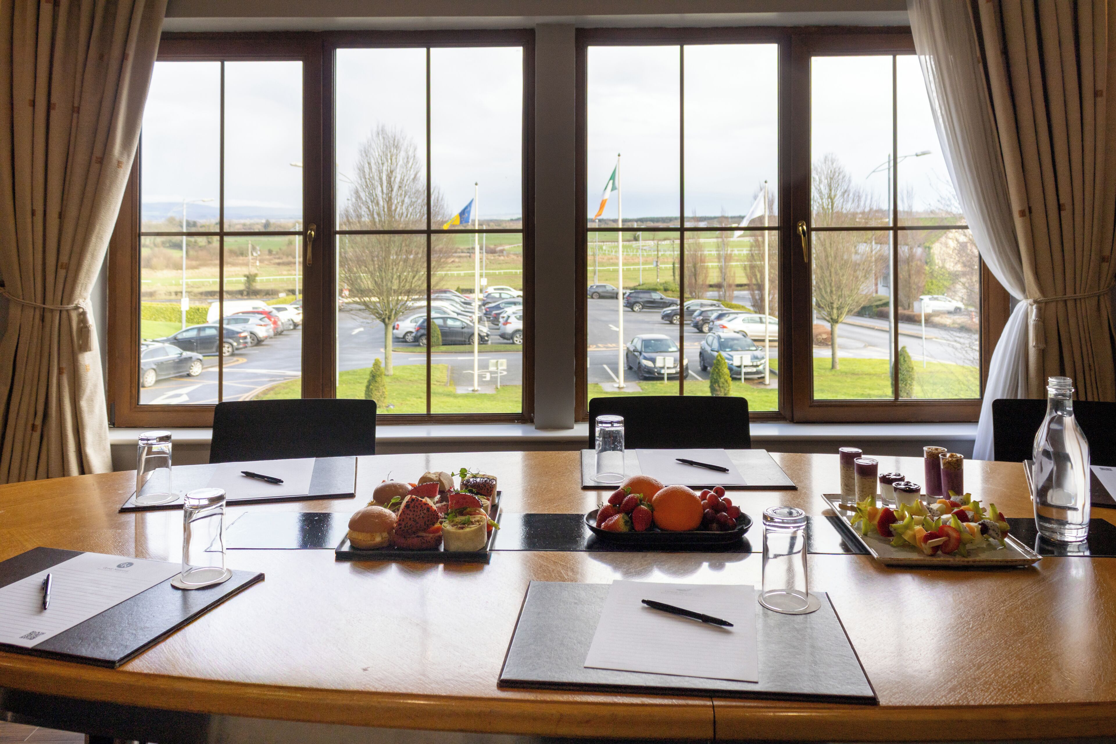 Une salle de conférence donnant sur un parking grâce à de grandes fenêtres, une table dressée avec de la nourriture et une bouteille d’eau.