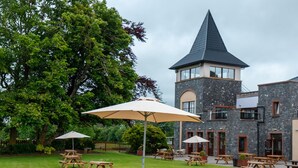 Un patio con mesas de picnic y sombrillas, un edificio de piedra con una torre y un árbol grande.