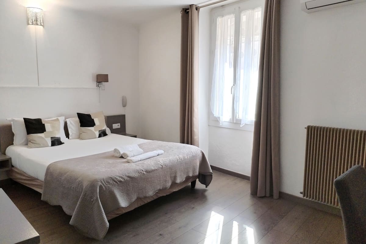Quarto de hotel com uma cama grande, mesa de cabeceira, escrivaninha, cadeira, janela com cortinas e luminária na parede.