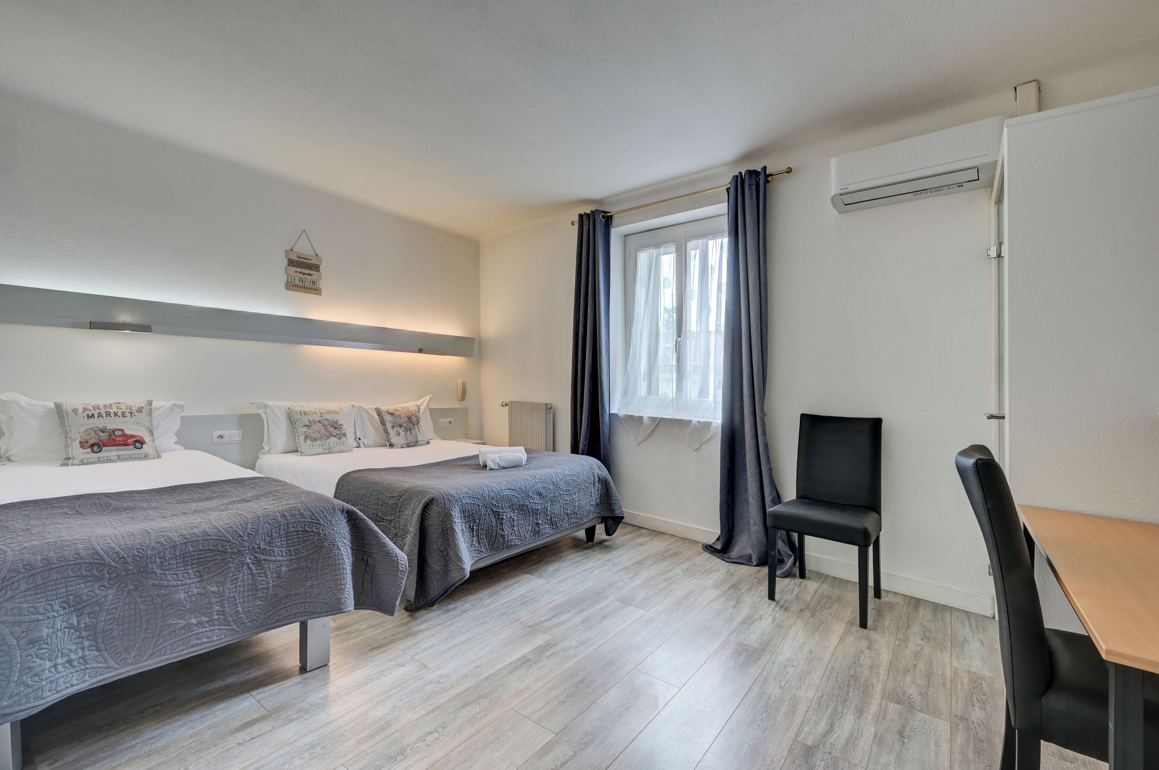 Ein Hotelzimmer mit zwei Betten, einem Schreibtisch, einem Stuhl, einem Fenster mit Vorhängen und einer Klimaanlage.