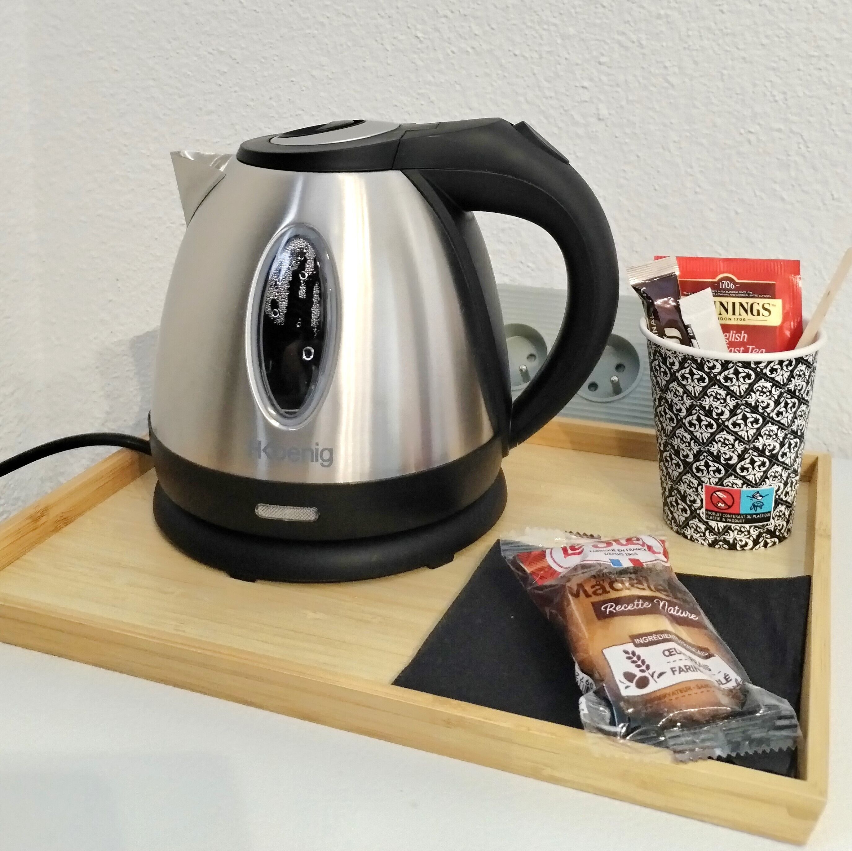Una tetera de acero inoxidable con un asa negra, una taza estampada y una bolsita de té sobre una bandeja de madera.