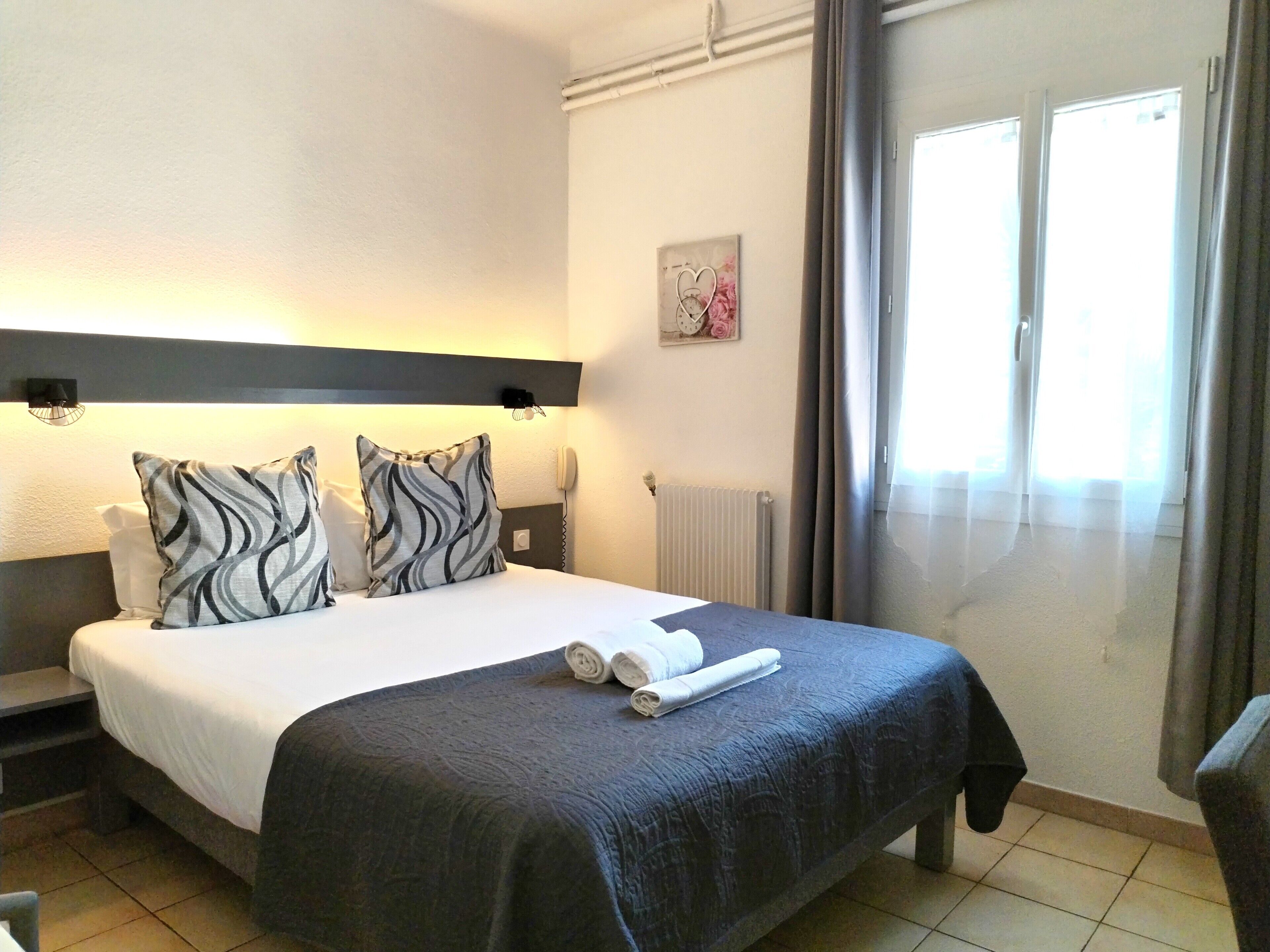 A hotel room with a bed, bedside tables, a telephone, a radiator, and a window with curtains.