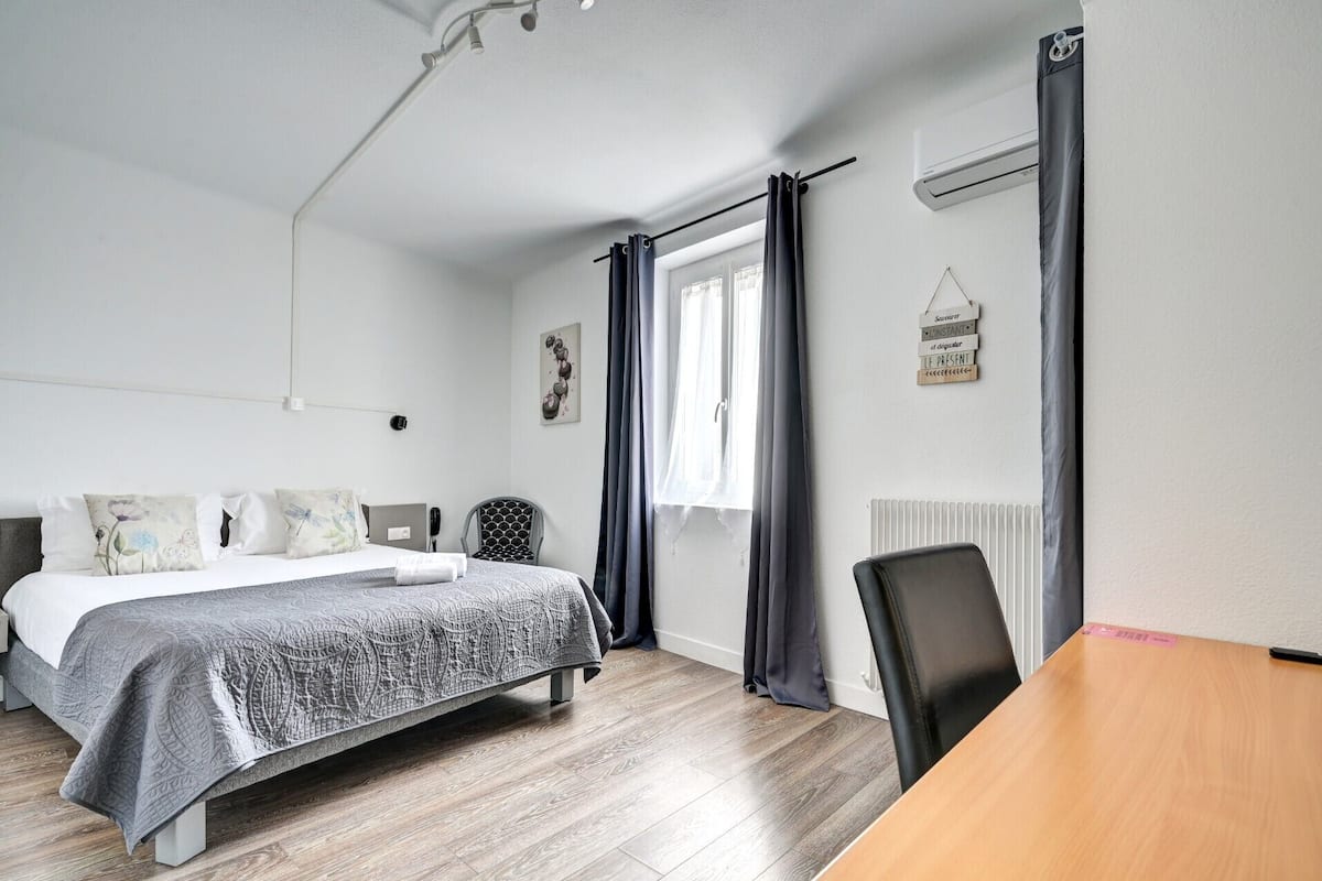 Quarto moderno com cama, escrivaninha e cadeira. Há uma janela com cortinas, radiador e ar condicionado fixado na parede.