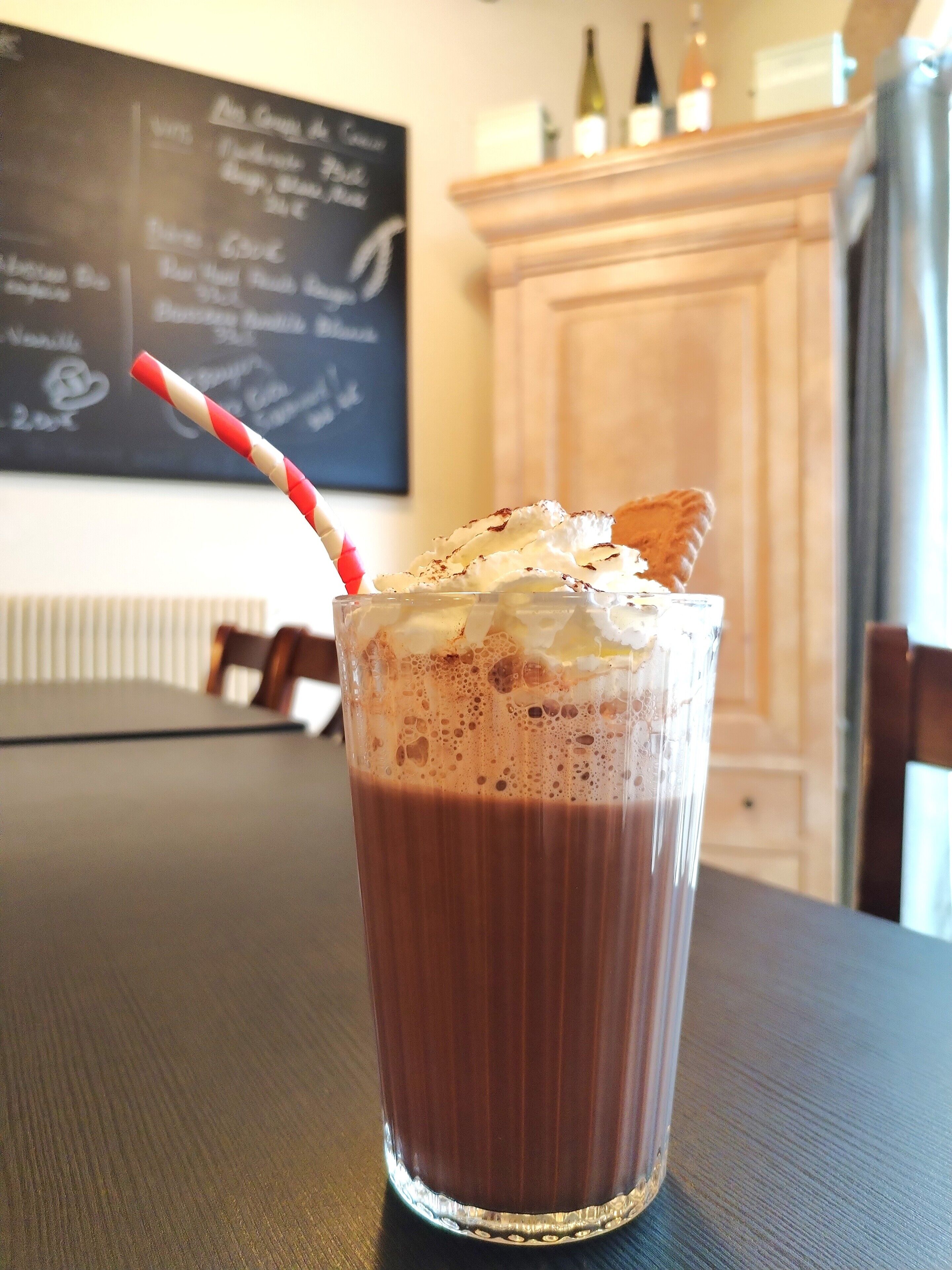 A glass of chocolate milkshake with whipped cream and a cookie, a red and white striped straw, and a blackboard menu in the background.