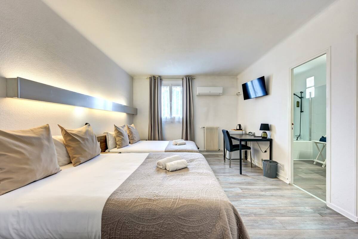 Quarto de hotel com duas camas, uma escrivaninha, uma cadeira, televisão e banheiro visível por uma porta de vidro.