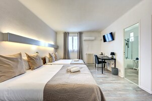 Habitación de hotel con dos camas, un escritorio, una silla, un televisor y un baño visible a través de una puerta de vidrio.