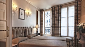 Ein Hotelzimmer mit zwei Betten, einer Sitzecke, einem Fenster mit Vorhängen, einer Kristallleuchtre und einem gerahmten Bild an der Wand.