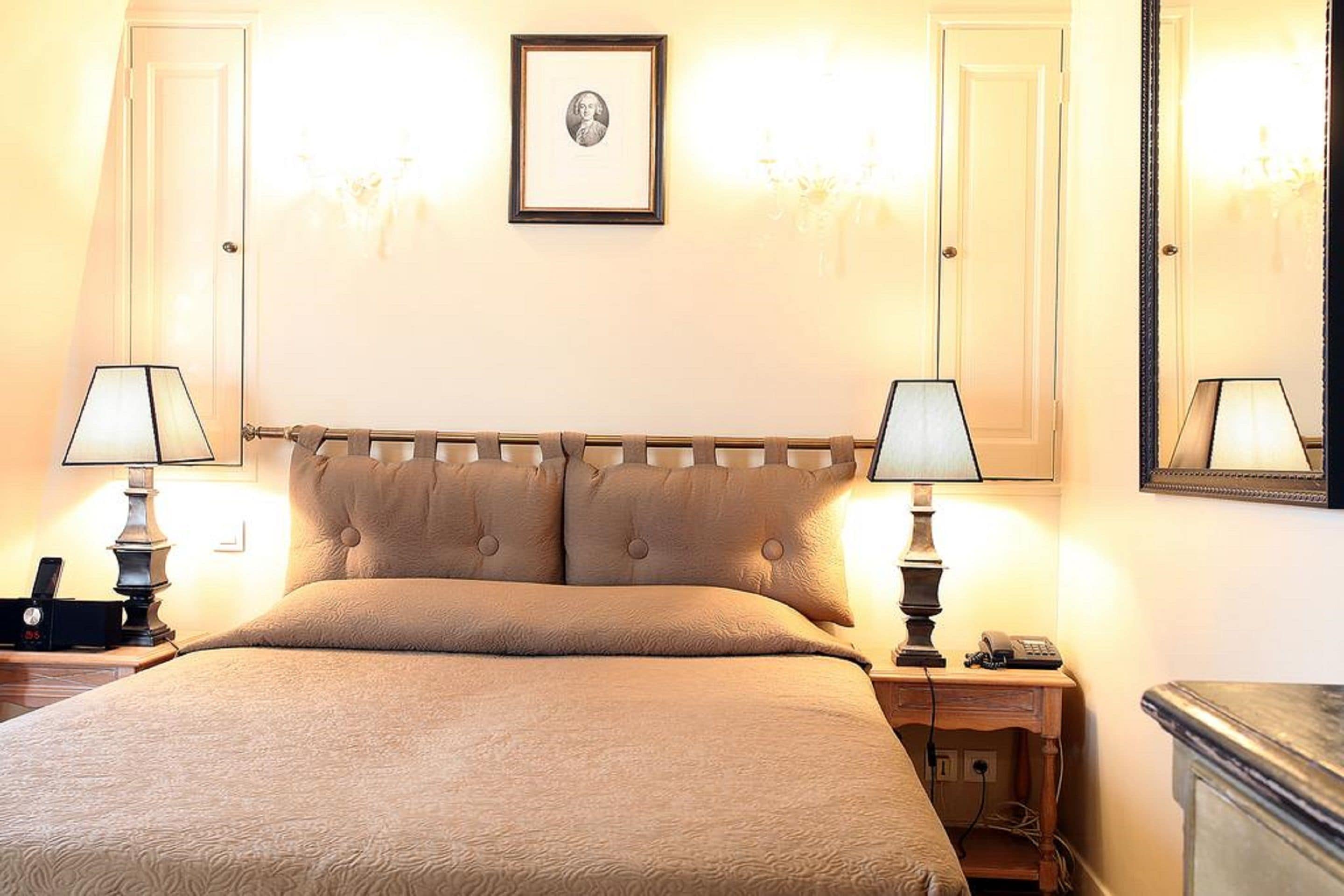 Una habitación de hotel con una cama grande, dos lámparas de noche, una mesita de noche, un cuadro enmarcado en la pared y un espejo.