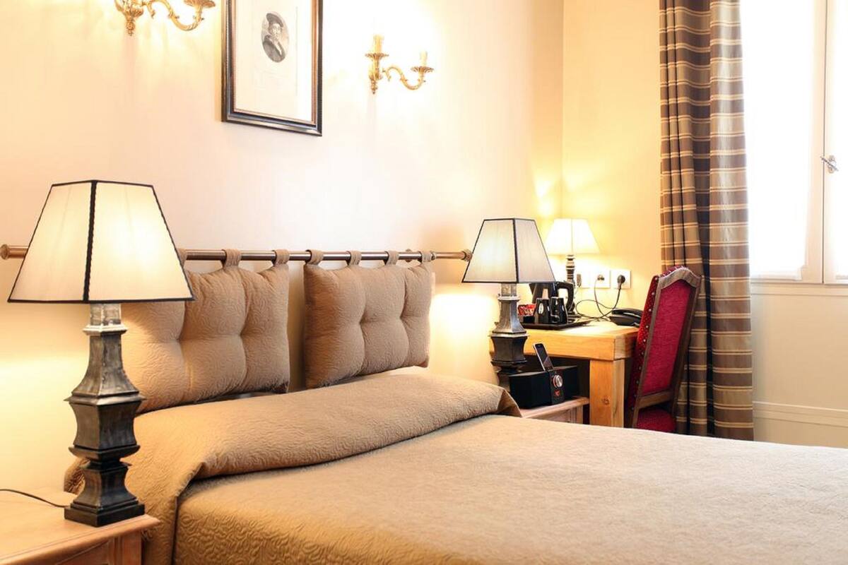A hotel room with a bed, two bedside lamps, a desk with a computer, a chair, and a window with curtains.
