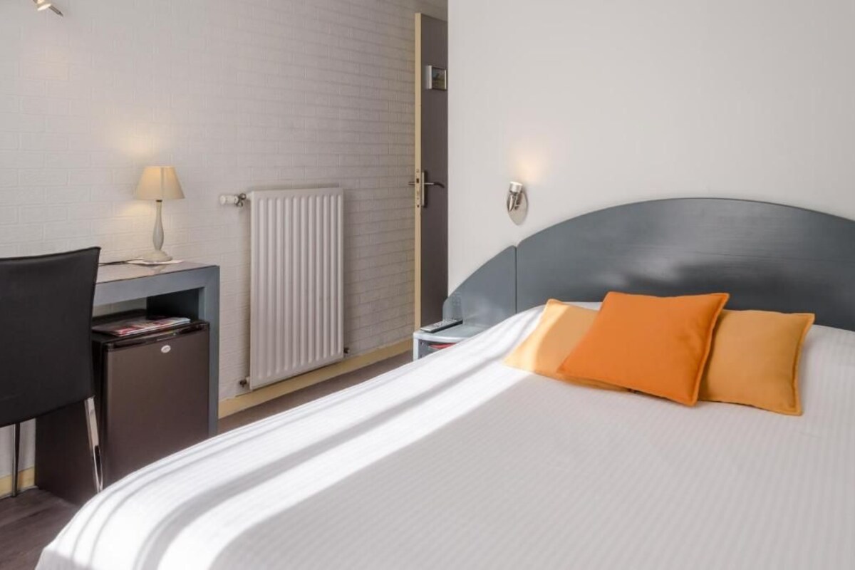 A hotel room with a bed, a desk with a lamp, a radiator, and a nightstand.