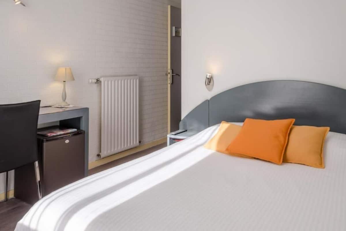 A hotel room with a bed, a desk with a lamp, a radiator, and a nightstand.