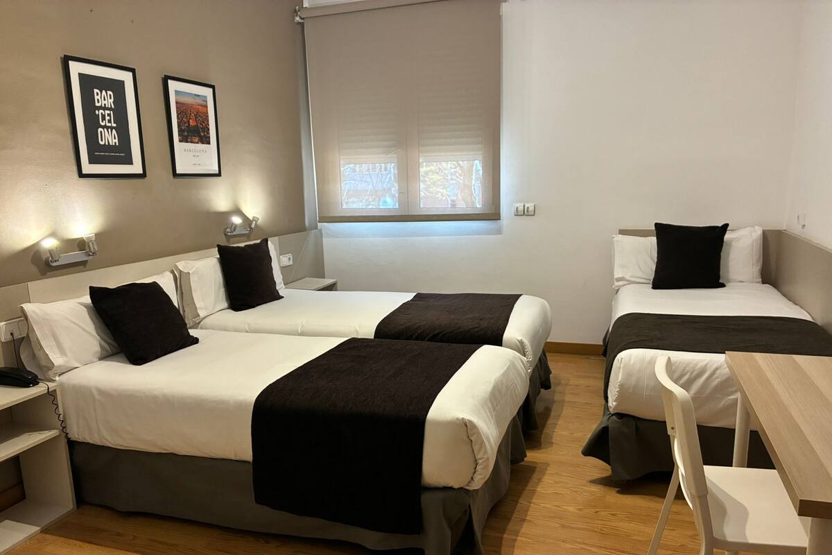 Habitación de hotel con dos camas, un escritorio, una silla, una ventana con persianas y dos cuadros en la pared.