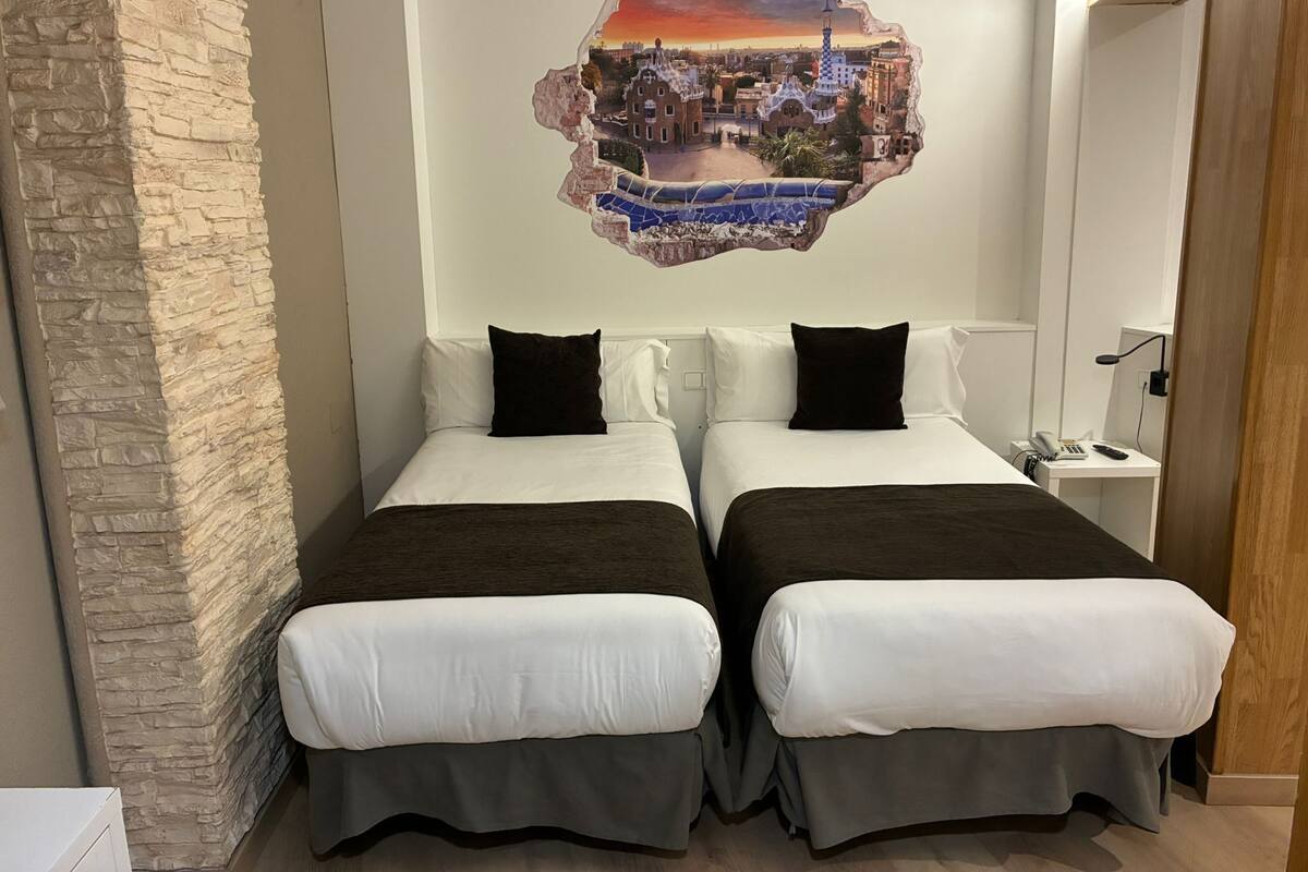 Habitación de hotel con dos camas, una pared de acento de piedra, un mural de un paisaje urbano y una mesita de noche pequeña con una lámpara.
