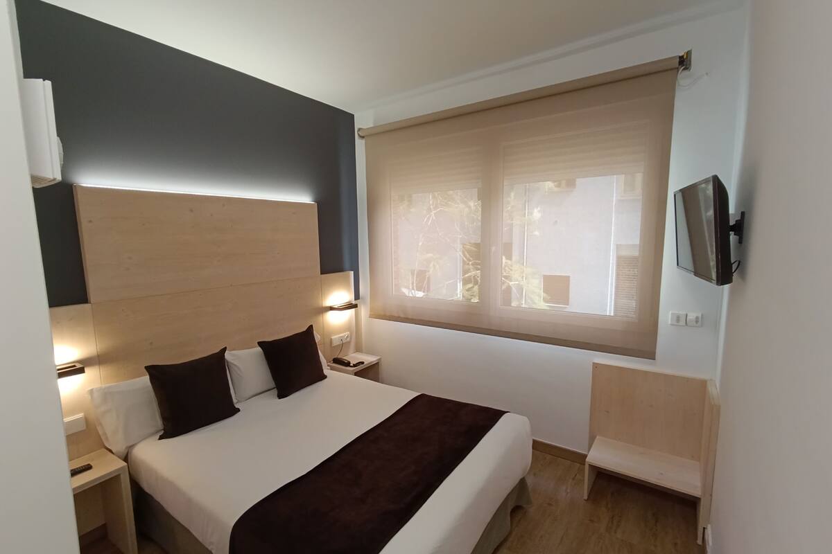 Una habitación de hotel con cama, mesitas de noche, televisión y ventana con persianas.