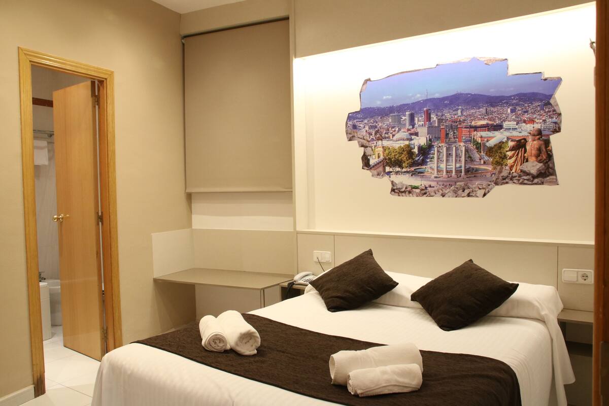 Habitación de hotel con una cama, dos almohadas, una colcha y un cuadro de un paisaje urbano en la pared.