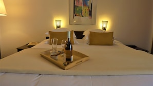 Habitación de hotel con una cama grande, dos mesitas de noche, una bandeja con una botella y vasos, y un cuadro en la pared.