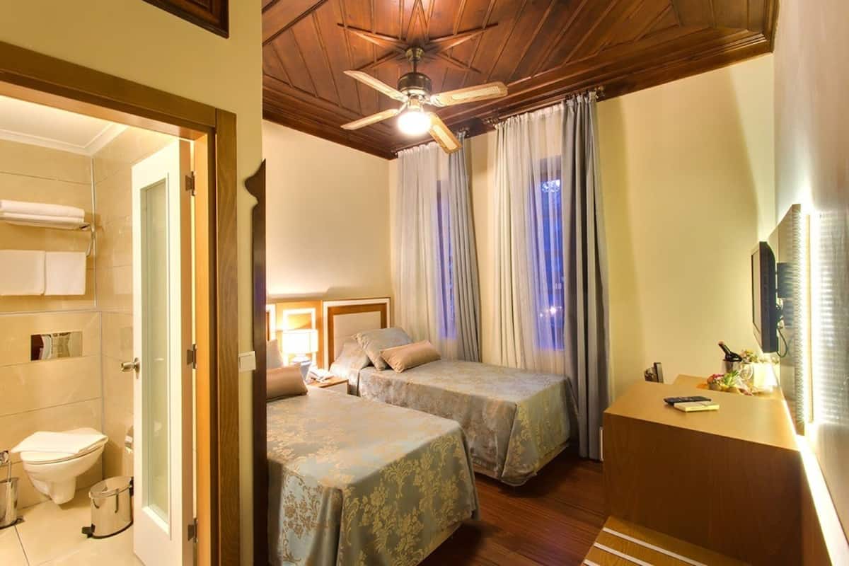 Ein Hotelzimmer mit zwei Betten, Holzboden, Deckenventilator, Badezimmer mit Toilette und Waschbecken sowie einem Schreibtisch mit Fernseher.
