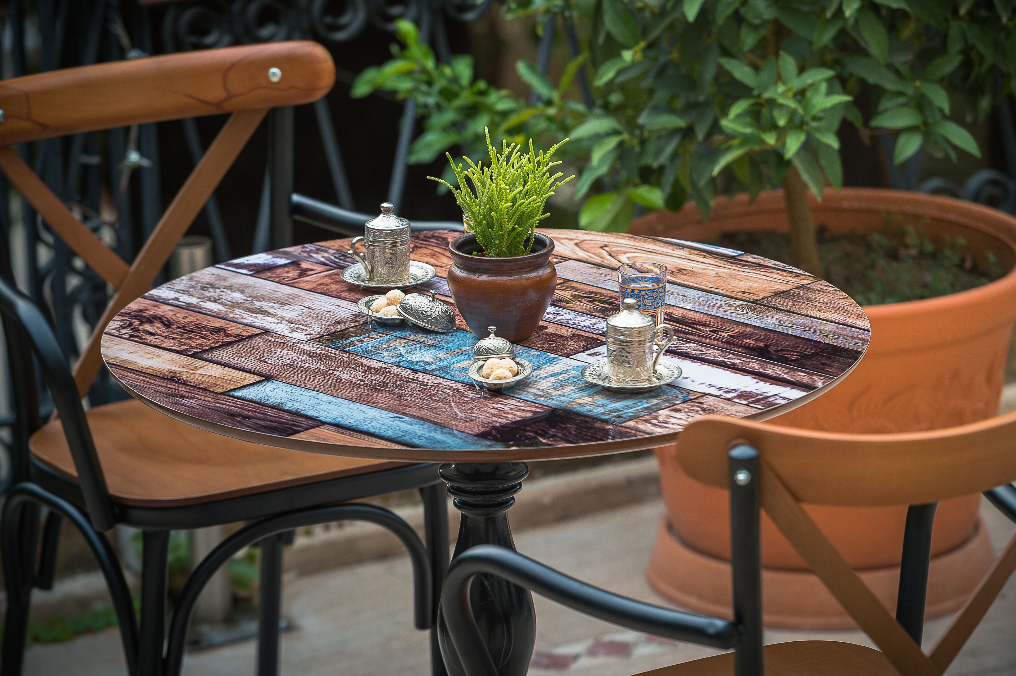 A round table with a rustic wooden top, surrounded by black metal chairs. A potted plant with green foliage is placed in the center.