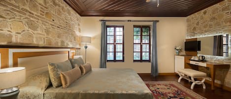 A bedroom with a stone wall, wooden ceiling, a bed with a patterned bedspread, a desk with a television, and a window with blue curtains.