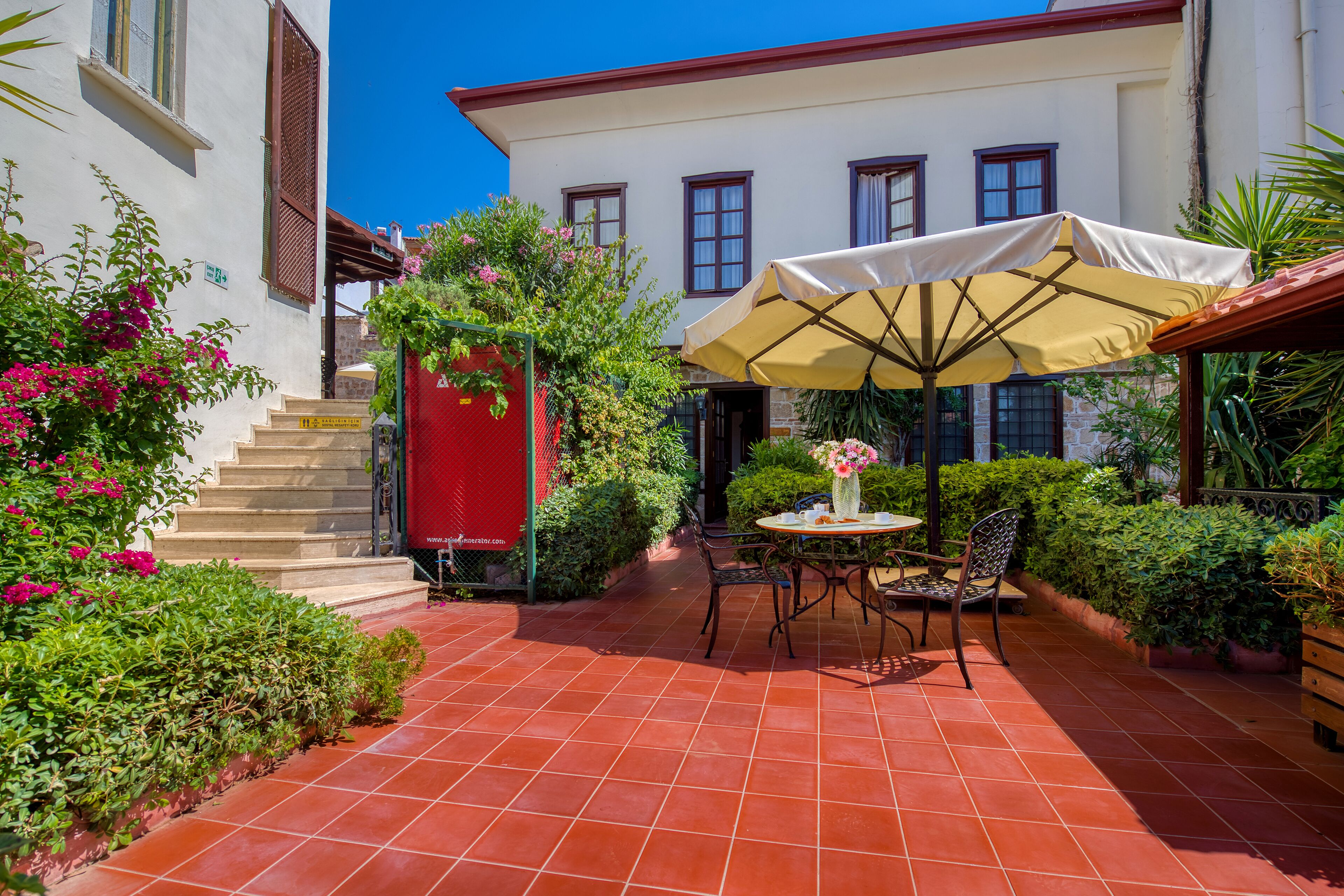 A patio area with a table and chairs, a red gate, and a building with multiple windows in the background.