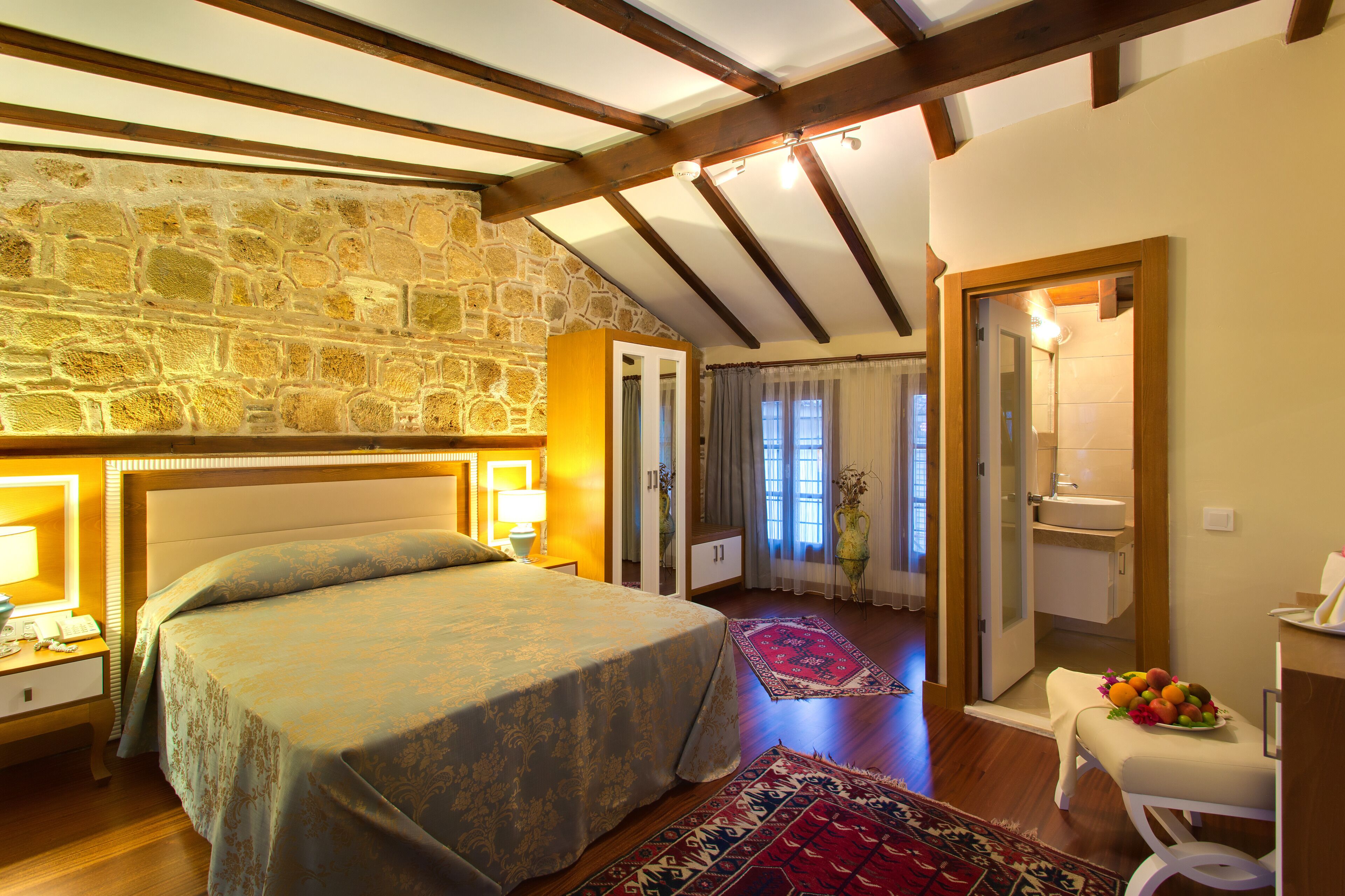 Ein Schlafzimmer mit Steinwand, Holzbalken, einem Bett mit gemustertem Überwurf, einem Nachttisch mit Lampe, einem durchgehenden Türspalt sichtbaren Badezimmer und einer Obstschale auf einer Bank.