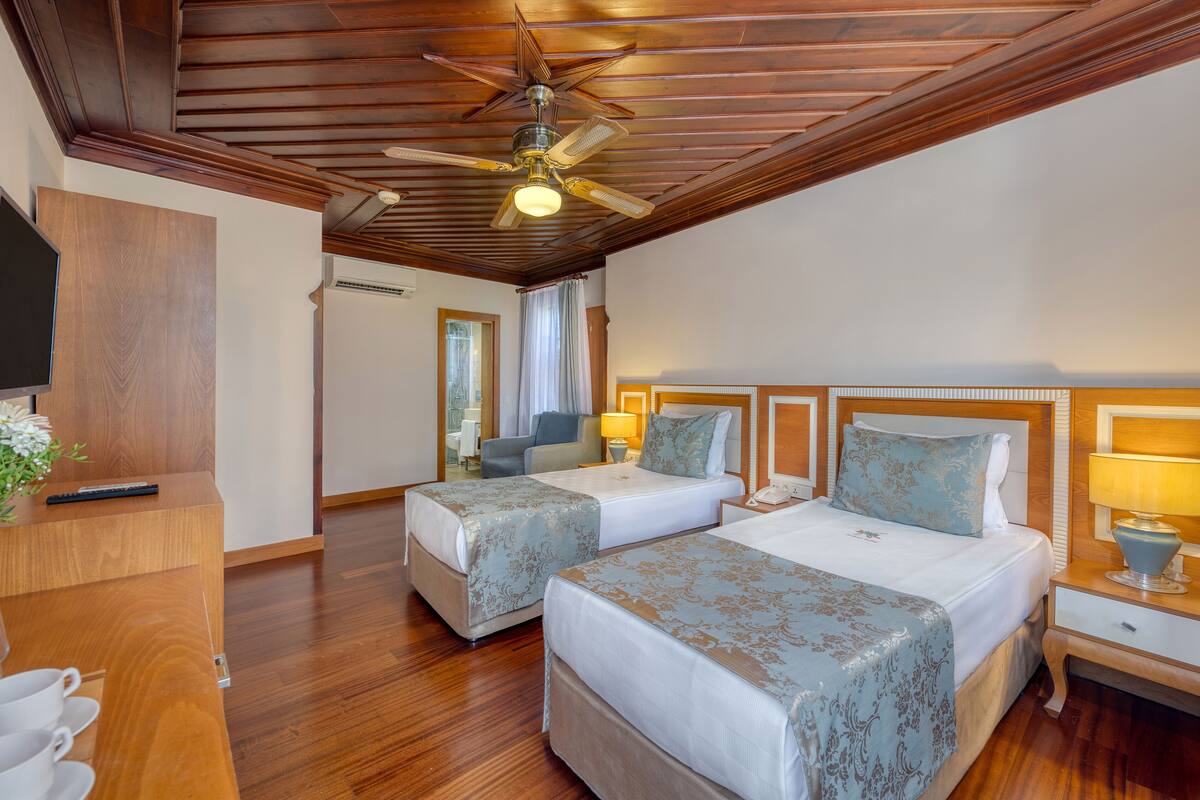 Ein Hotelzimmer mit zwei Betten, Holzboden, Deckenventilator, Fernseher und einem Schreibtisch mit Lampe.