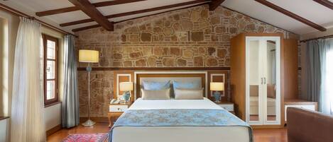 A bedroom with a stone wall, wooden beams, a large bed, bedside lamps, a nightstand, a wardrobe, and a red patterned rug.