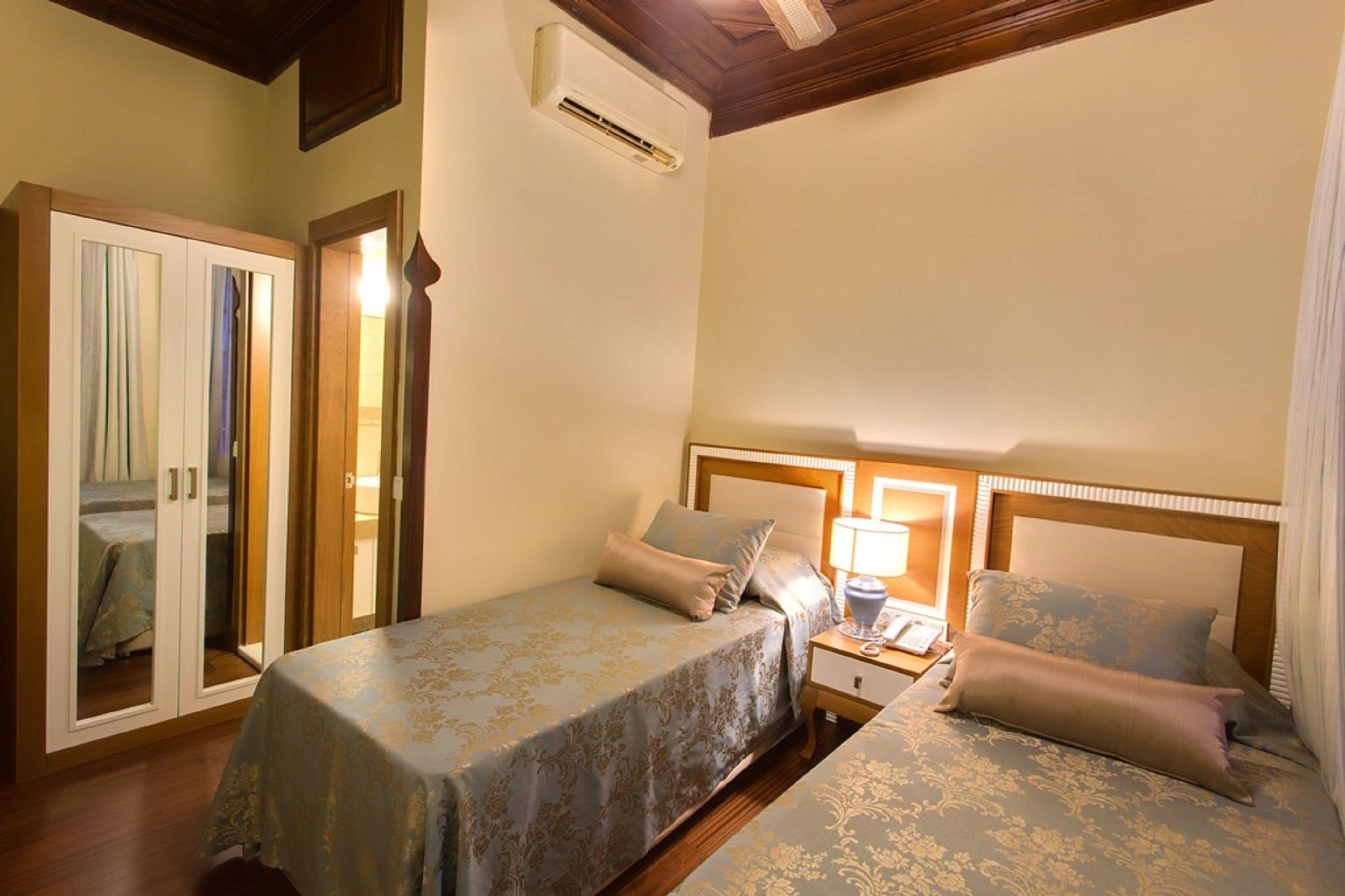 A hotel room with two beds, a wardrobe, an air conditioning unit, and a bedside lamp.